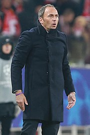 A white man in a black suit, watching a football match and giving orders to the players.