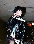 一個戴著黑假髮，穿著黑白鄉間的夾克對著麥克風唱歌的女性。