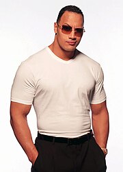 Vue de face du catcheur The Rock. Il est vêtu d'un T-shirt blanc et d'un pantalon noir.