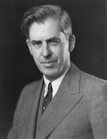 Black-and-white image of the head and shoulders of man about fifty with upswept hair, wearing a gray suit and a dark tie