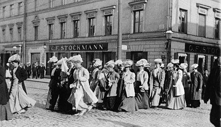 Seitliche Schwarzweißfotografie einer Frauengruppe, die jeweils zu viert aufgereiht eine Straße überquert. Sie tragen lange Kleider und breite Hüte. Im Hintergrund ist eine Häuserfassade mit Geschäft zu sehen.