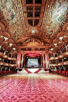 A large room with red floor and red and gold decoration to the walls. There is a stage at the back of the picture with seating areas to the sides.