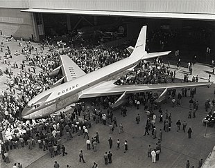 Vue plongeante sur un avion, devant un hangar d'assemblage, avec une foule de personnes rassemblées tout autour ; la photo est en noir et blanc.