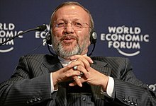 Portrait d'un homme, portant des lunettes et un casque audio, croisant les mains, assis devant un fond indiquant World Economic Forum.