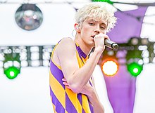 Un jeune homme blond et mince portant un sweat sans manches chante sur une scène, un micro à la main, en regardant directement dans l'objectif du photographe