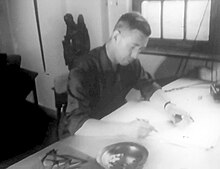 Photographie en noir et blanc d'un homme aux cheveux courts tenant un crayon de la main droite, légèrement penché en avant