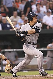 A man in a grey baseball uniform and blue batting helmet swings a baseball bat standing at home plate.