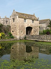 A two-storey stone building with a vehicular arch through the centre. It has a stone roof and the gables at each end have stone ball finials. In front of the gatehouse is a stone bridge across a wide moat that has water plants growing on it.