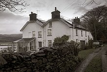 Photographie en noir et blanc de deux maisons accolées aux murs blancs, celle de gauche garnie d'une petite véranda, avec au premier plan et sur la gauche, un mur de pierres bordant le chemin d'accès ; à l'arrière plan on entrevoit un plan d'eau.