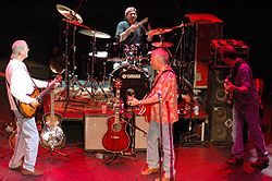 Three men playing guitars and one playing drums, performing together on a stage