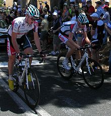 A pair of cyclists on the road, wearing matching white jerseys with blue and red trim. A car is partly visible behind them, and numerous spectators are on the roadside.