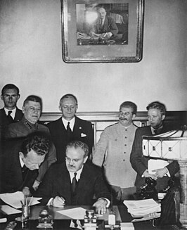 Ondertekening van het Molotov-Ribbentroppact. De Duitse minister van Buitenlandse Zaken Joachim von Ribbentrop staat achter Molotov die tekent. Jozef Stalin glimlacht op de achtergrond rechts van hem.