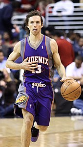 A white basketball player, wearing a purple jersey with a word "Phoenix" and the number "13" written in the front, dribbles the ball