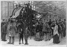 An engraving of a large printing press twice as tall as a person. A crowd of onlookers is gathered around it.