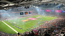 A large soccer field seen from the upper stands of a stadium near the rafters; the view is partially obstructed by a railing and other fans. On the field, the flags of Spain and England and a large banner with the Women's World Cup trophy are held for display.