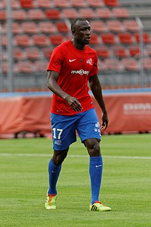 Photographie d'un homme sur un terrain de football avec un maillot rouge, un short bleu, des chaussettes bleues et des chaussures de football jaunes. En arrière-plan, une tribune