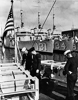 Американские и британские моряки стоят у глубинных бомб. На заднем плане — американские эсминцы типа «Викс» переданные по договору «эсминцы в обмен на базы»