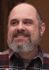 A 51 year-old man with a grey beard smiling to the left of the camera