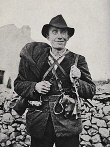 Photo noir et blanc mal définie d’un homme armé, portant un chapeau, souriant, devant un mur en pierres.