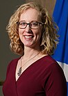 A white woman with curly blonde hair. She has glasses on. Behind her is a Scottish flag.