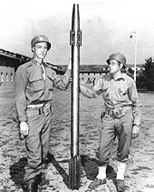 Two soldiers in US Army uniforms hold between them a long, slim projectile that is somewhat taller than them, with a finned tail