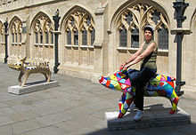 Tow pig statues in front of yellow stone building with arched windows. The nearest status is multi coloured band being ridden by person.