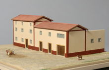 Photographie de la maquette d'une maison gallo-romaine. Les murs sont beige et rouge, le toit imite des tuiles. Il y a deux étages au bâtiment principal, et les portes sont en bois.