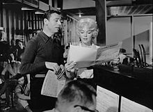 Monroe y Montand de pie junto a un piano en un entorno tipo estudio y mirando partituras.