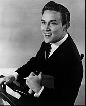 A dark-haired man wearing a dark jacket and tie, sitting in a director's chair