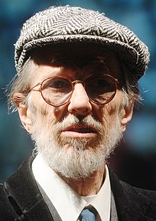 An elderly man with a white beard, round glasses, a beret-like hat, a dark vest, and a necktie. He faces down right, looking into an open book.