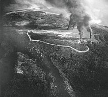 Black and white aerial photo of an open area in a jungle with several columns of smoke rising from it