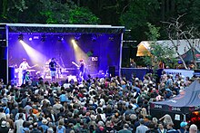 Blau beleuchtete Festivalbühne vor Waldhintergrund. Auf der Bühne sieht man eine Band spielen, davor viele Hinterköpfe und Rücken von Zuschauern und in deren Mitte das schwarze Zelt der Festivaltechnik.