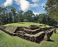 A low, wide structure with stepped sides, with the top covered in low grass. A dense treeline forms the backdrop to the structure. The right hand side of the structure has several standing stones before it, with a local guide standing by one of them.