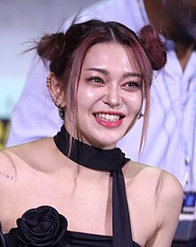 Image of AleXa smiling; she has two hair buns and she is wearing a black dress and many earrings