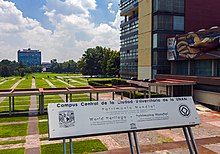 Un cartel metálico prateado nos soportes que se afasta da cámara que di "World Heritage Site" en español e inglés, con texto de apoio. Detrás hai un edificio cun mural, unha pasarela cuberta e un gran quad con edificios ao lonxe