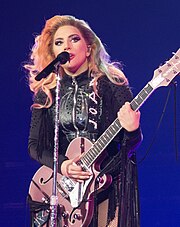 Lady Gaga standing behind a microphone stand with a pink guitar in her hands, wearing black leather fringe.