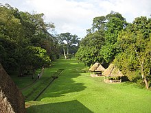Un espacio abierto y llano de hierba, rodeado de árboles, con unos pocos techos de paja que cubren los monumentos y al lado izquierda un canal de drenaje y un sendero