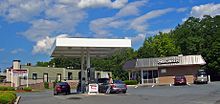 A one-story brownish building with flat, flared roof and the script "Stewart's" in white on the roof on the right. In the center, in the foreground, are several gas pumps under a small canopy. At left is a maroon-and-white sign with another script "Stewart's" and gas prices.