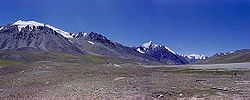 Some tall, blue-grey mountains rise out of brown soil below a deep blue sky