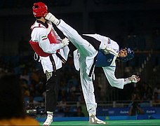 photographie de deux hommes en kimono blanc en train de se battre. Ils portent des équipement de protection bleu pour l'un, et rouge pour l'autre. Celui portant un équipement bleu est en train d'assener un coup de pied au visage de celui portant un équipement rouge.