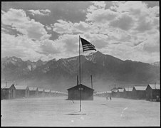 photo d'un camp. Au loin de hautes montagnes enneigées sont visibles. Au centre un mat en haut duquel un drapeau américain est visible et domine deux rangés de bâtiments présents de part et d'autre. Le terre-plein central est balayé par le vent et de nombreuses poussières sont soulevées.