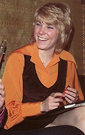 A blonde woman wearing an orange blouse and a black vest, smiling broadly