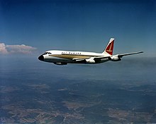 Photo couleur d'un avion en vol vu de trois quart face. Le ciel est presque totalement dégagé et un relief vallonné se situe sous l'appareil, qui se trouve à haute altitude.