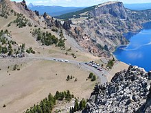 Vue plongeante depuis des rochers sur une route et un parking dominant le lac cerné par des reliefs déchiquetés.