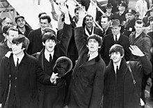 Fotografía en blanco y negro de los Beatles saludando frente a una multitud con un par de escaleras de avión en el fondo