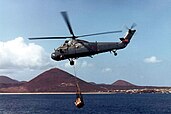 A helicopter flies over the water with a load slung below. In the background is an island with a series of pyramid-shaped peaks.