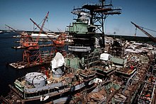 In the background a large shipyard can be seen, with cranes and dockyard warehouses. In the foreground a massive steel superstructure can be seen, decked out in scaffolding with tarps covering sensitive equipment and wires across the superstructure in all directions.