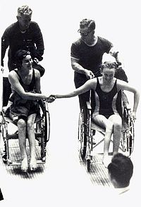 Australian Paralympic Team members Daphne Ceeney (now Hilton) and Elizabeth Edmondson shake hands after Edmondson won gold and Ceeney silver in the 50m prone swimming event at the 1964 Tokyo Paralympic Games. They are pushed by Kevin Betts (Ceeney) and 'Johnno' Johnston (Edmondson)