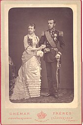 Photo en noir et blanc d'un couple de fiancés, à gauche, la jeune fille, esquissant un léger sourire, porte une longue robe claire aux jupes bouffantes et enlace de ses mains le bras de son fiancé portant un uniforme à épaulettes et un sabre, son visage est impassible.