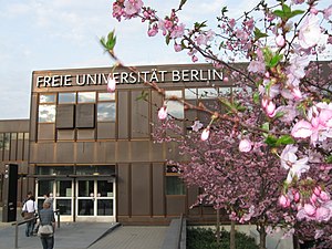 Frontale Farbfotografie eines dunkelbraunen Gebäudes mit der Aufschrift „Freie Universität Berlin“. Das zweigeschossige Gebäude wird rechts teilweise von einem Kirschblütenzweig verdeckt.